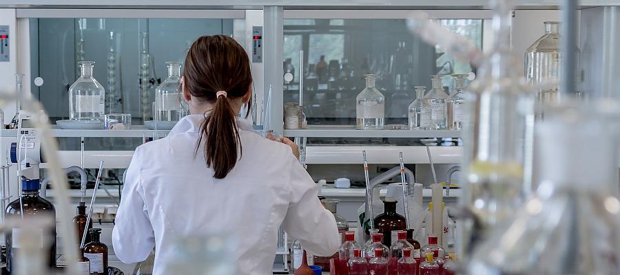 Zdjęcie przedstawiające kobietę w białym fartuchu, która pracuje w laboratorium. Wokół niej znajduje się szklany sprzęt laboratoryjny (butelki, kolby, próbówki, zlewki itp.) 